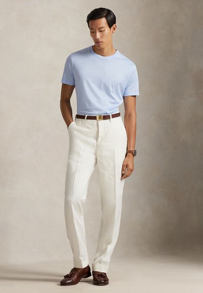 Άνδρας που φορά φωτιστό γαλάζιο πουκάμισο με κοντά μανίκια, λευκό παντελόνι με καφέ ζώνη, καφέ μοκασίνια με φούντες και ρολόι χειρός, στέκεται μπροστά σε μπεζ φόντο.