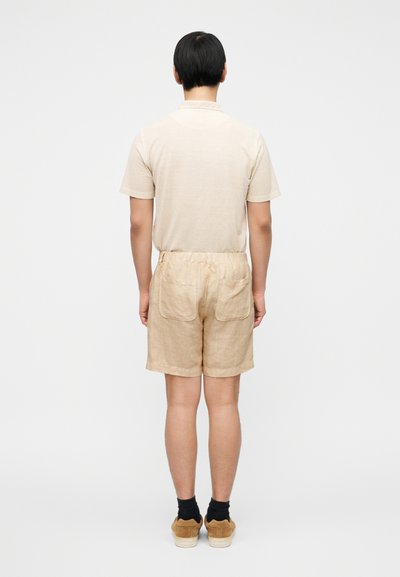 Μπεζ λινό πόλο πουκάμισο και ταιριαστά σορτς με ελαστική μέση και δύο πίσω τσέπες, φορεμένα με μαύρες κάλτσες και καφέ παπούτσια.