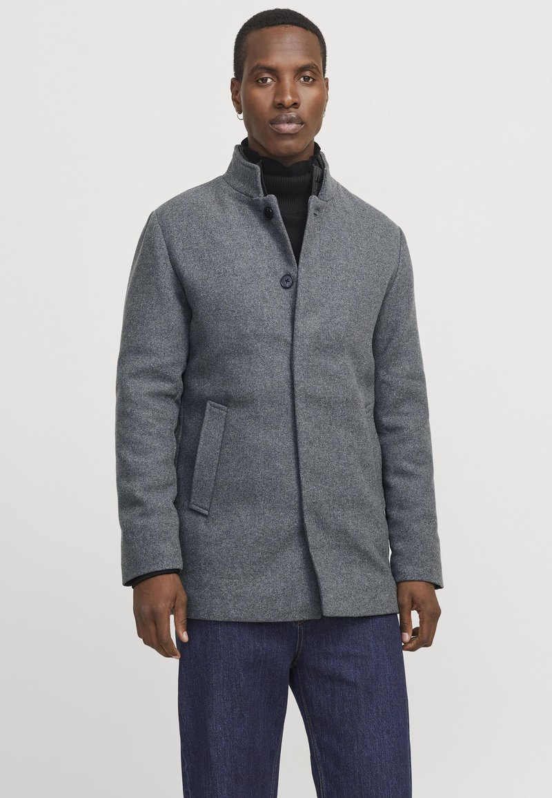 Homme portant un manteau grison et un pull à col roulé noir, se tenant contre un fond blanc uni.