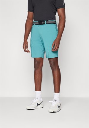 Mies, jolla on turkoosit shortsit, musta vyö, musta paita, valkoiset Nike-tennarit ja valkoiset matalat sukat, seisoo yksinkertaisen vaalean taustan edessä.