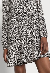 Zwarte jurk met witte bloemenprint; lange mouwen, gathered manchet en een relaxte pasvorm. Materiaal lijkt licht en vloeiend.