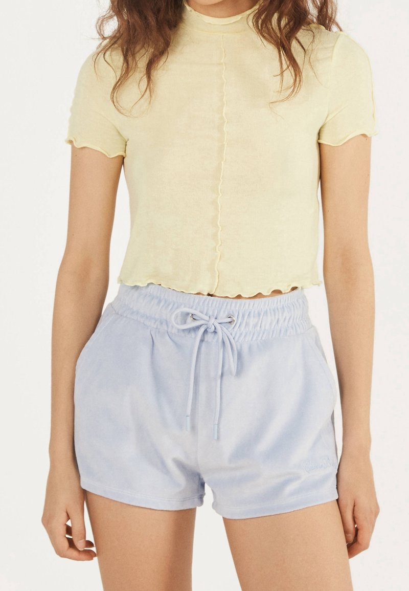Femme portant un crop top jaune clair à manches courtes et un short bleu clair à cordon, debout avec les bras détendus le long du corps.