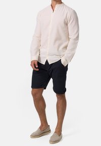 Chemise à manches longues, beige clair, avec un col mandarin, associée à un short bleu marine foncé et des espadrilles gris clair.