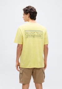 Άνδρας που φοράει κίτρινη μπλούζα Patagonia με λογότυπο βουνών στην πλάτη και καφέ cargo σορτς, στέκεται μπροστά σε απλό φωτεινό φόντο.