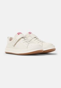 Sneakers bianchi in pelle con una texture liscia, dotati di una fascia in velcro e dettagli rossi del logo, poggiati su una suola in gomma marrone chiaro.