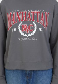 Sudadera gris con texto rojo que dice "MANHATTAN", "NYC" y "La ciudad que nunca duerme", con un diseño circular de laurel.