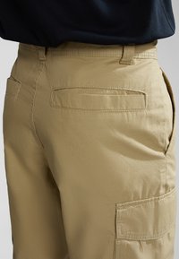 Pantaloni cargo khaki con una texture liscia, tasche laterali e posteriori, e una vita aderente. Cuciture visibili lungo le cuciture.