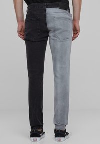 Jeans de denim con un diseño dividido, una pierna negra y la otra gris claro. Bolsillos traseros visibles; la tela tiene una textura suave.