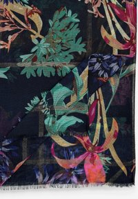 Tissu sombre avec motif botanique coloré, plié à un coin, montrant des bords frangés et des motifs floraux superposés en bleu sarcelle, rose et orange.