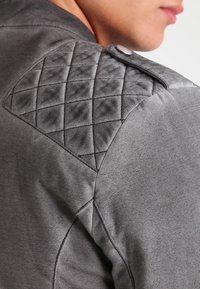 Giacca grigia con dettaglio trapuntato sulle spalle, texture morbida e un pulsante decorativo sull'epaulette. Primo piano della costruzione del capo.