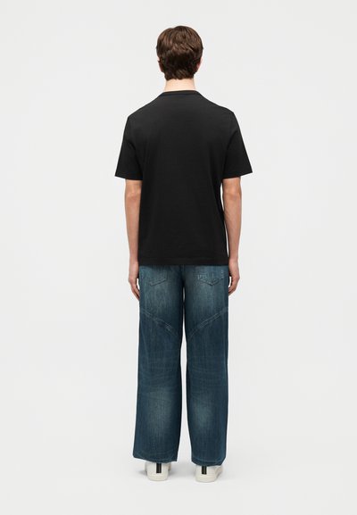 Νεαρό άτομο στέκεται με την πλάτη, φορώντας απλό μαύρο κοντομάνικο μπλουζάκι, μπλε παντελόνι με φαρδιά μπατζάκια και λευκά αθλητικά παπούτσια με μαύρες λωρίδες.