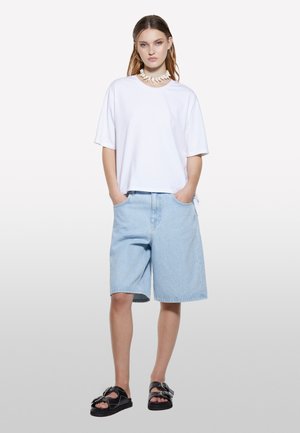 T-shirt en coton blanc avec une coupe décontractée, associée à un short en denim bleu clair. Des sandales noires avec des éléments décoratifs complètent le look.