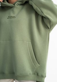 Jasnozielona bluza z kapturem wykonana z miękkiego materiału, z przednią kieszenią i wyhaftowanym w ciemniejszym zielonym napisem "BĄDŹ WYJĄTKOWY BĄDŹ SOBĄ".