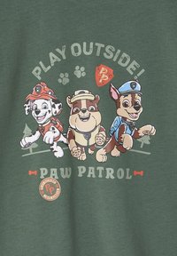 Grünes T-Shirt mit cartoonartigen Hunden in Rettungsanzügen, mit dem Text "DRAUSSEN SPIELEN!" oben und "PAW PATROL" unten, mit Pfotenabdrücken.