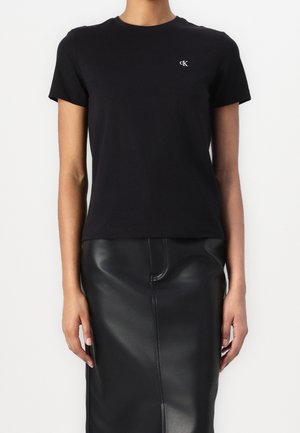 Black cotton t-shirt with short sleeves and a round neckline, featuring a small white logo on the chest. Paired with a black leather skirt.