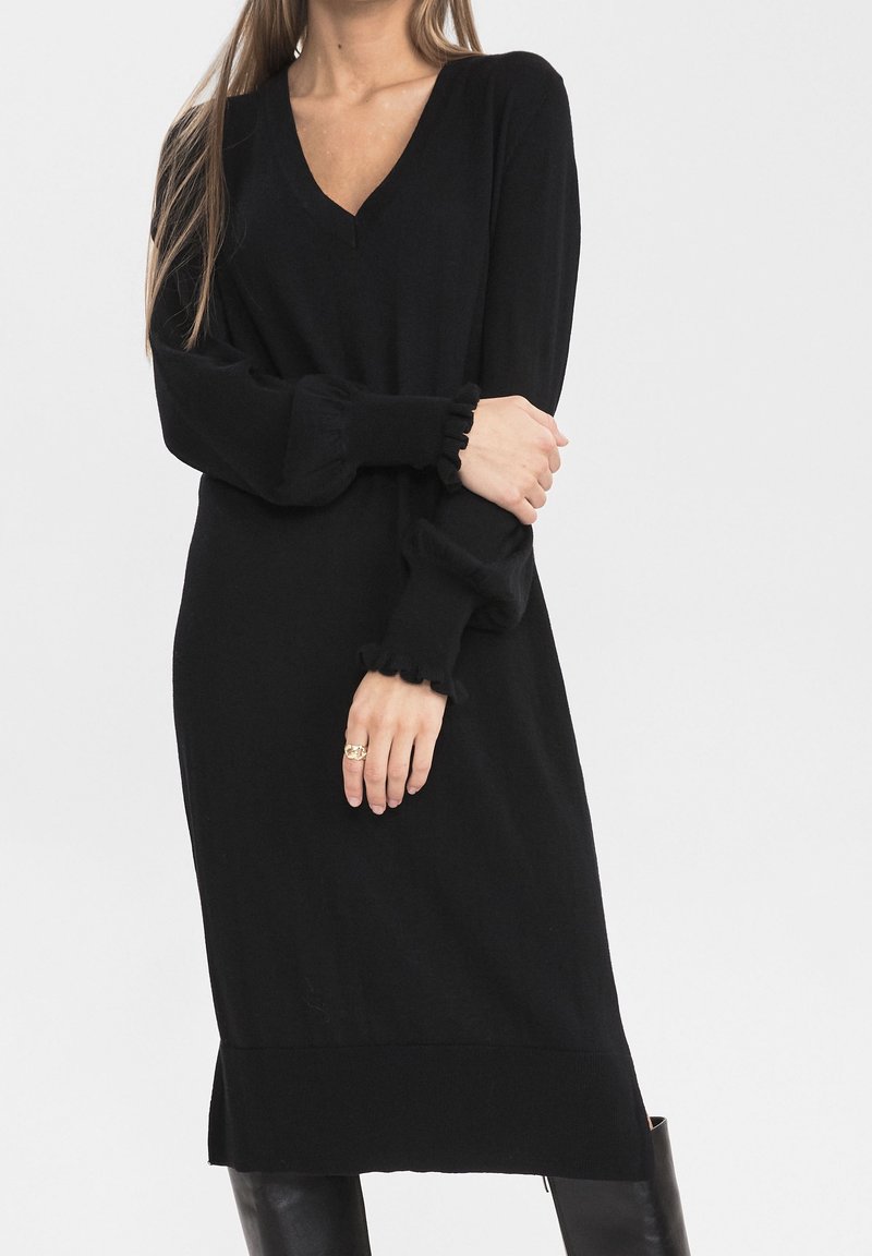 Robe en maille noire avec un col en V, des manches longues ornées de poignets à volants et une silhouette droite se terminant au-dessus du genou. Texture lisse.
