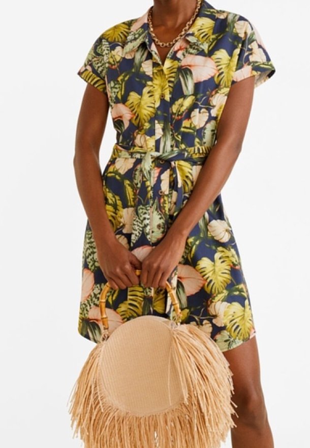 Femme portant une robe boutonnée à manches courtes avec un imprimé coloré de feuilles tropicales, tenant un sac à main rond en paille avec des détails à franges.
