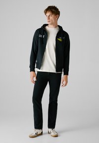 Crna zip-up dukserica s detaljima loga, slojevita preko kreme majice, u kombinaciji s tamnim trapericama i bijelim tenisicama s crnim akcentima.
