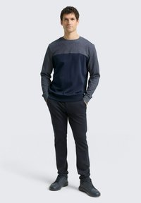 Granatowa i szara koszula z długim rękawem, z jednolitą górną częścią, w zestawieniu z ciemnymi spodniami o dopasowanym kroju i ciemnymi trampkami. Gładkie, teksturowane materiały.