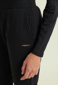 Pantalones deportivos negros hechos de tela suave con un bolsillo lateral texturizado, que presentan un logo dorado "FREDDY" y una cintura acanalada.