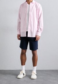 Světle růžová košile s knoflíky vyrobená z bavlny, v kombinaci s námořnickými šortkami. Model má na sobě bílé tenisky a bílé ponožky, pod košilí má jednoduché bílé tričko.