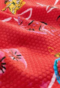 Piros textúrájú szövet, színes virágmintákkal, hatszögletű mintákkal és kontrasztos rózsaszín, sárga és zöld hímzéssel.