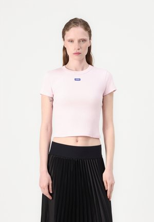 Lichtroze, cropped t-shirt met korte mouwen, geribbeld textuur en een klein blauw logo patch op de borst, gecombineerd met een zwarte geplooide rok.