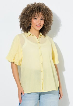 Frau mit lockigem Haar, die eine hellgelbe Bluse mit Knöpfen und hellblaue Jeans vor weißem Hintergrund trägt.