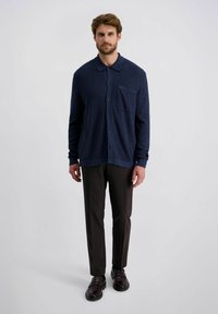 Marineblaue Hemdjacke mit Tasche, aus strukturiertem Stoff, kombiniert mit dunkelbraunen maßgeschneiderten Hosen und glänzenden schwarzen Schuhen.