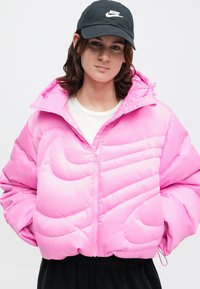 Φουσκωτό ροζ μπουφάν με ψηλό γιακά, μπροστινό φερμουάρ, διαγώνιες ραφές και ελαστικές μανσέτες. Φορεμένο με λευκό πουκάμισο από κάτω.