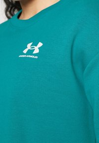 Turkusowy sweatshirt wykonany z miękkiego materiału, z okrągłym dekoltem, ściągaczami w rękawach i białym logo Under Armour na klatce piersiowej.