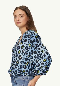 Jasnoniebieska bluzka w czarno-zielony wzór w panterkę, luźny krój, szerokie rękawy i dekolt w kształcie litery V.