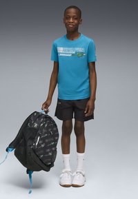 Turquoise T-shirt met korte mouwen met een "SUPER PUMA" print, zwarte shorts, witte schoenen, en een zwarte rugzak met groene accenten die in de hand wordt gehouden.