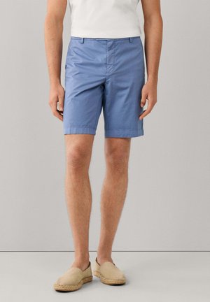 Man draagt lichtblauwe knielange short, wit T-shirt en beige instapschoenen, staande tegen een effen grijze achtergrond.