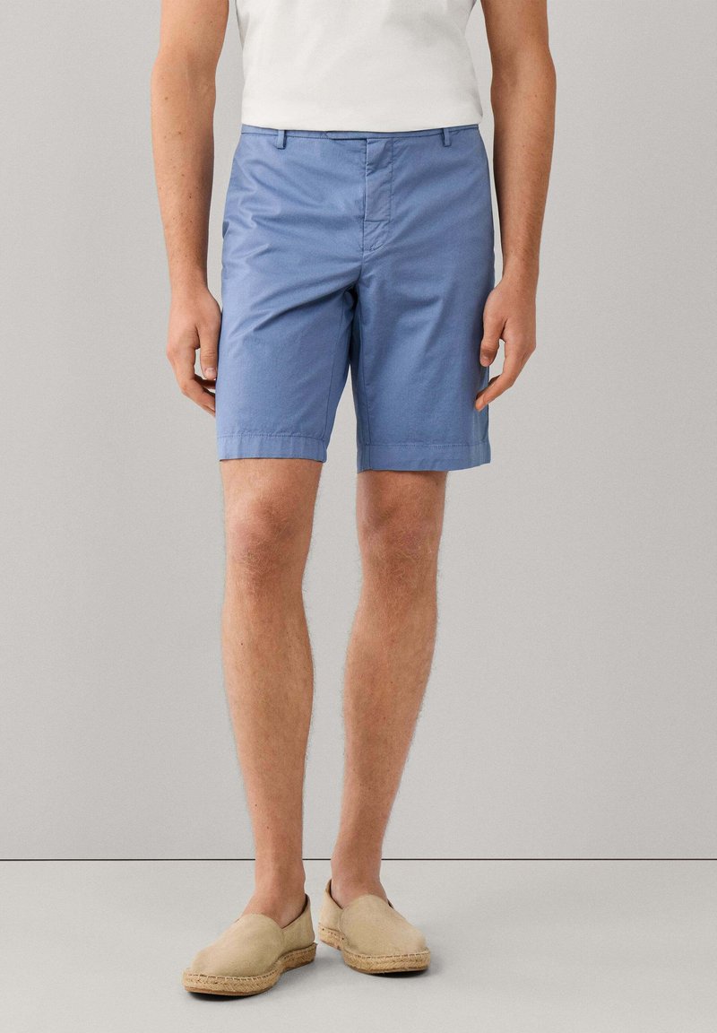 Hombre con pantalones cortos azul claro hasta la rodilla, camiseta blanca y zapatos beige sin cordones, de pie contra un fondo gris liso.