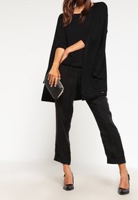 Svart öppen cardigan, figurnära svart topp, lösa svarta byxor och svarta högklackade skor. Håller en liten silverklutch.