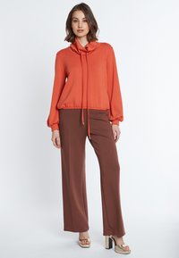 Oranges Oberteil mit Kragen und langen Ärmeln, kombiniert mit braunen weiten Hosen und goldenen Blockabsatzsandalen. Weicher Stoff, lockere Passform und modernes Design.