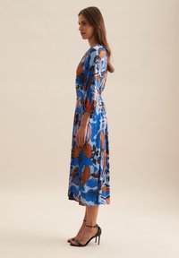 Blaues Blumenkleid mit orangefarbenen und schwarzen Akzenten, knielang, mit puffärmel und tailliertem Schnitt. Das Model trägt schwarze Absatzsandalen.