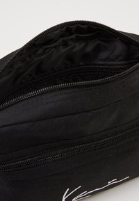 Czarna torba na suwak z zamknięciem, charakteryzująca się gładką teksturą i białym logo. Wewnątrz znajduje się jedna komora z czarnym wykończeniem.