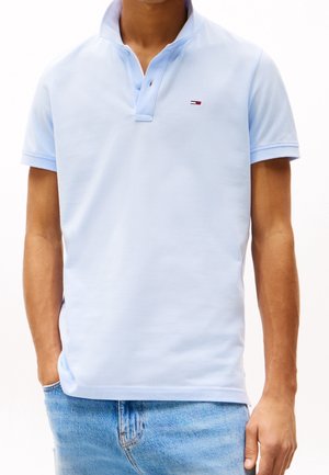 Mand iført en lyseblå kortærmet polo med en lille rød, hvid og blå rektangulær logo, kombineret med blå jeans.