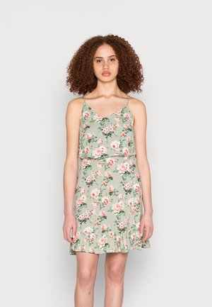 Vestido floral em tecido verde claro com alças finas. Apresenta uma bainha com volantes e um padrão de flores rosa e brancas sobre um fundo de folhas.