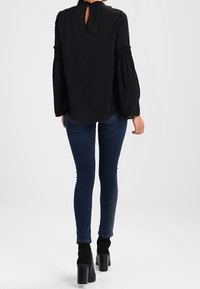 Schwarze Langarmbluse mit gerafften Bündchen und Schlüsselloch-Detail im Rücken, kombiniert mit dunkelblauen, schmal geschnittenen Jeans und schwarzen Stiefeletten.