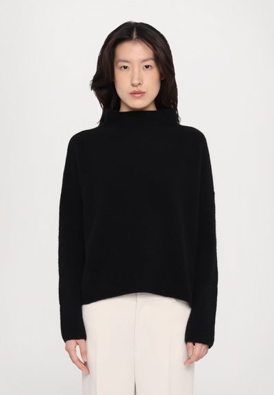 Pull noir à col haut en tissu doux et texturé. Doté de manches longues et d'une coupe décontractée, associé à un pantalon de couleur claire.