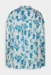 Chemise à motif floral avec une base crème clair et des designs bleus. Manches longues, col, et une coupe ample avec un tissu texturé.