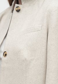 Ελαφρύ μπεζ μάλλινο παλτό με σχέδιο ψαροκόκαλο, κουμπιά καφέ χελώνας και τσέπη στο στήθος, φορεμένο πάνω από λευκή ριμπ σκουφάκια.