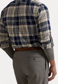 Mies, jolla on sinivihreä ja beige ruudullinen pitkähihainen paita harmaan neuleen päällä, harmaat housut ja ruskea nahkavyö, näkymä takaa.