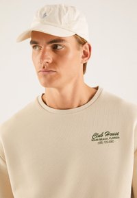 Beige sweatshirt med rund hals, med grønn tekst "Club House, Miami Beach, Florida," kombinert med en hvit caps med logo.