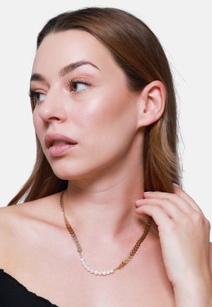 Collana d'oro con elementi a forma di foglia e perle disposte in una linea curva. Modella che indossa un top nero senza spalle.