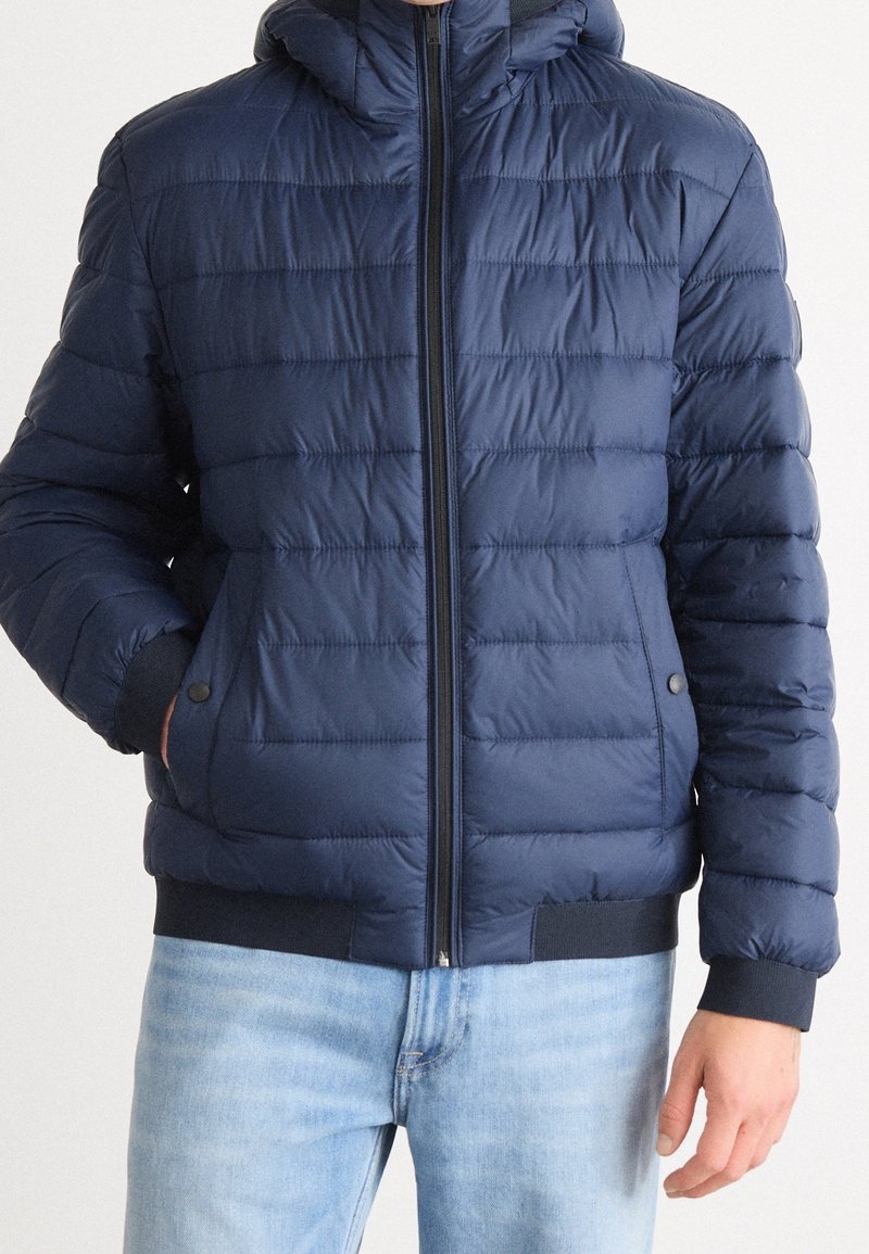Homme portant une veste matelassée bleu marine avec fermeture éclair et poches latérales, associée à un jean bleu clair, debout avec une main dans la poche.