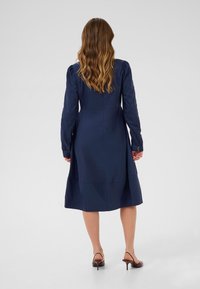 Tamsiai mėlyna suknelė su ilgomis rankovėmis, prigludusia juosmeniu ir platėjančia sijonu. Yra mygtukais užsegamos rankovės ir lygus, tekstūruotas audinys.